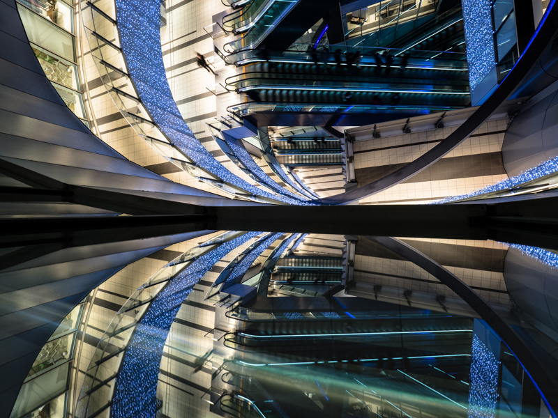 映り込みを生かしたシンメトリー構図で日常的な風景を非日常的に切り取った1枚。1/2秒のスローシャッターでぶらした歩行者がアクセントだ。のぞき込むように手持ち撮影する不安定な状況でも強力な手ぶれ補正の効果で背景はシャープに写り、静と動を対比できた。<br><span class="fnt-85">オリンパス OM-D E-M1 Mark Ⅱ / M.ZUIKO DIGITAL ED 7-14mm F2.8 PRO /7mm（14mm相当）/マニュアル露出（F9、1/2秒）/ ISO 200</span>