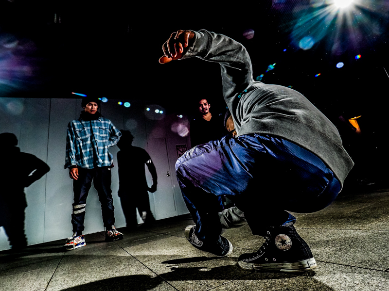 バリアングルモニターと軽量でグリップのあるコンパクトなカメラ。フットワークの軽さが功を奏し、後ろに立つ二人が画に入り込む位置に一瞬で立ちまわることができた。その結果、手前のダンサーとの意味合いを持たせることに成功した。<br><span class="fnt-85">オリンパス OM-D E-M1 Mark Ⅱ / M.ZUIKO DIGITAL ED 7-14mm F2.8 PRO / 7mm（14mm相当）/ マニュアル露出（F5.6、1/125秒）/ ISO 400 / ダンサー: MATSU</span>