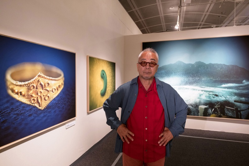 藤原新也。金の指輪、碧玉製勾玉、沖ノ島の荒海の写真の前で。