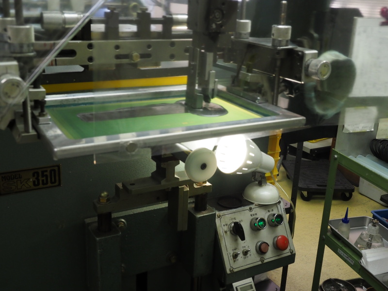 シルク印刷の機械。ライトで照らされた白い円盤（写真中央部）にフィルター枠をセットし、上からインクをのせる。