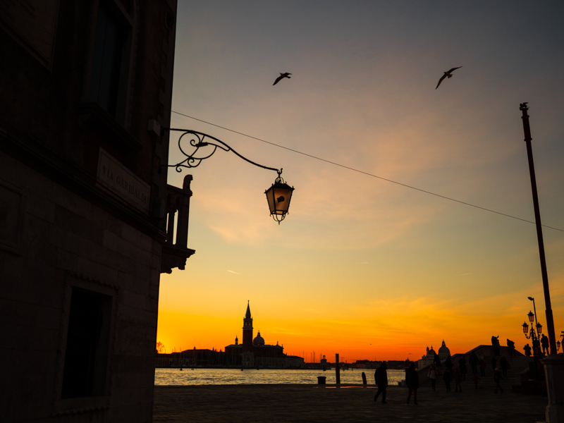 海をはさんだサンジョルジョ島方向へと沈んだ夕日の残照。ベネチア本島の街並みから明かりがつく直前の薄暗い時間に、F1.2の明るさで寝床へと帰る鳥たちのシルエットを止めた。<br><span class="fnt-85">OLYMPUS OM-D E-M1 Mark II / M.ZUIKO DIGITAL ED 17mm F1.2 PRO / 17mm（34mm相当） / 絞り優先AE（F1.2、1/6,000秒、+0.5EV） / ISO 200</span>