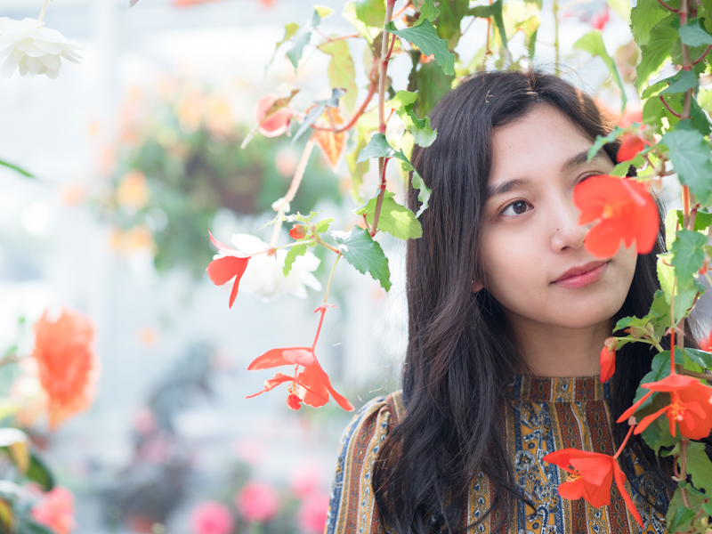 鮮やかな赤い花が印象的だったので、モデルにその花と戯れてもらった。背景のにじむボケが、情景を物語りつつも被写体の存在感を引き立てている。<br><span class="fnt-85">OLYMPUS OM-D E-M1 Mark II / 45mm（90mm相当） / マニュアル露出（F1.6、1/800秒） / ISO 400</span>