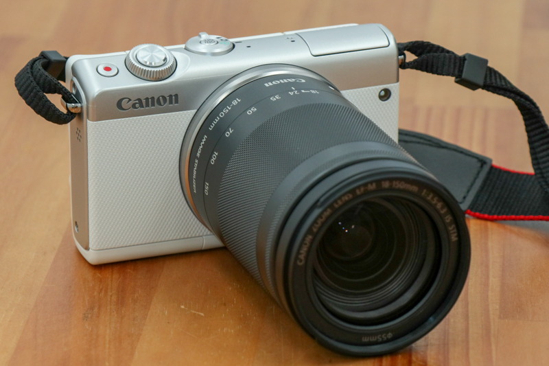 交換レンズ「EF-M 18-150mm F3.5-6.3 IS STM」を装着した状態のミラーレスカメラ「EOS M100」。