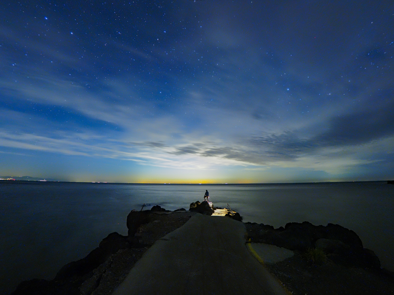 北の方角を望む海岸線は、東京の街明かりを受けてオレンジ色に輝く。長時間露光で波の動きを平滑化し、インターバル撮影で自分を入れた（撮影：北山輝泰）<br><span class="fnt-85">OLYMPUS OM-D E-M1X / M.ZUIKO DIGITAL ED 8mm F1.8 Fisheye PRO / 8mm（16mm相当） / マニュアル露出（F1.8、25秒） / ISO 1600 / WB：3,000K</span>