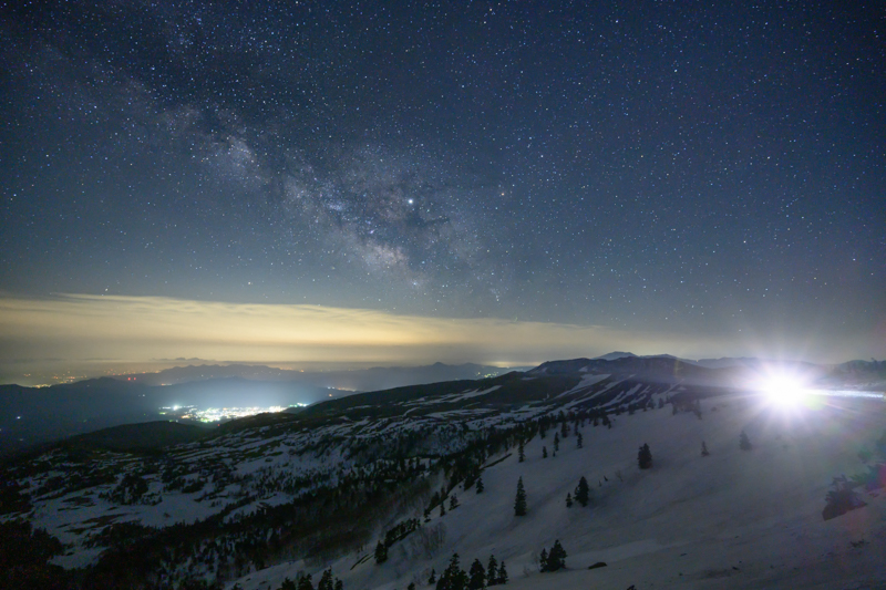 標高2,172mから望む春の雪山と天の川を14mmの超広角で狙う。嫌われがちな車のヘッドライトも捉え方によっては良いアクセントになる。開放F値でも画像周辺部のコマ収差が少なく、Z 7の優れた高感度耐性によって、レンズの描写力を存分に引き出している。<br><span class="fnt-85">Z 7 / NIKKOR Z 14-30mm f/4 S / 14mm / マニュアル露出（F4・20秒） / ISO  4000</span>