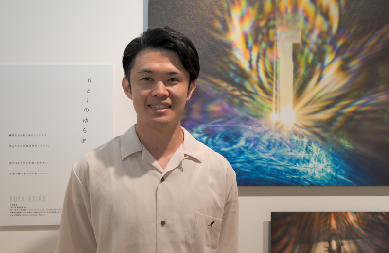 小池裕也さん。エキシビジョンの作品「0と1のゆらぎ」の前で。ピンホールを使用して光を捉えたという作品で、作品パネルに掲出されているQRコードから動画作品の世界へも誘う展示となっていた。展示作品は静止した光だが、動画では揺れ動く光が鑑賞できる仕組みだ