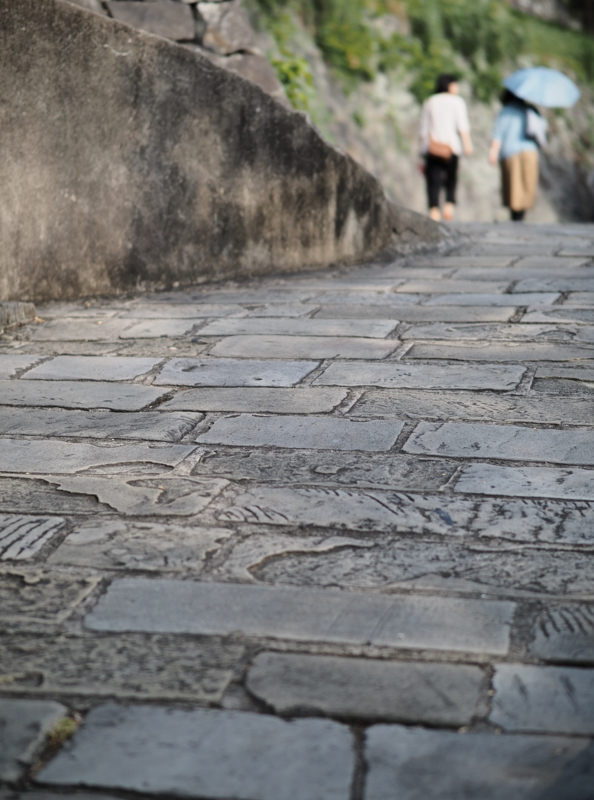 江戸時代、丘の上に住むオランダ人たちが使っていたオランダ坂と呼ばれる石畳の道。坂の途中にカメラを構え、すり減った石を見つめ、昔の人が行き交う様子を想像しながら、絞りを開放F1.2にしてタイミングよく背景に人をぼかした（撮影：斎藤巧一郎）。<br><span class="fnt-85">OLYMPUS OM-D E-M5 Mark III / M.ZUIKO DIGITAL ED 45mm F1.2 PRO / 45mm（90mm相当） / 絞り優先AE（F1.2、1/4,000秒、-0.3EV） / ISO 200 / WB：オート</span>