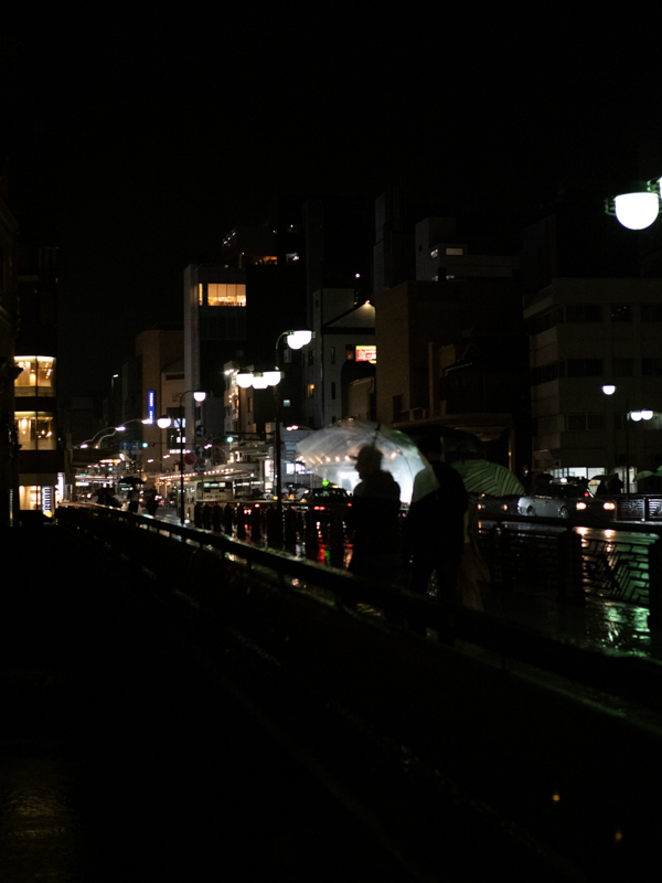 古くから交通の要所である四条大橋。傘越しに覗くシルエットが印象的で思わずシャッターを切った。E-M1 Mark II ・F1.2PROレンズの組み合わせであればAF低輝度限界は-6EVである。雨天・夜間こそ好ましいシチュエーションだ（撮影：中条望）。<br><span class="fnt-85">OLYMPUS OM-D E-M1 Mark II / M.ZUIKO DIGITAL ED 25mm F1.2 PRO / 25mm（50mm相当） / 絞り優先AE（F1.8、1/500秒、±0EV） / ISO 3200 / WB：オート</span>