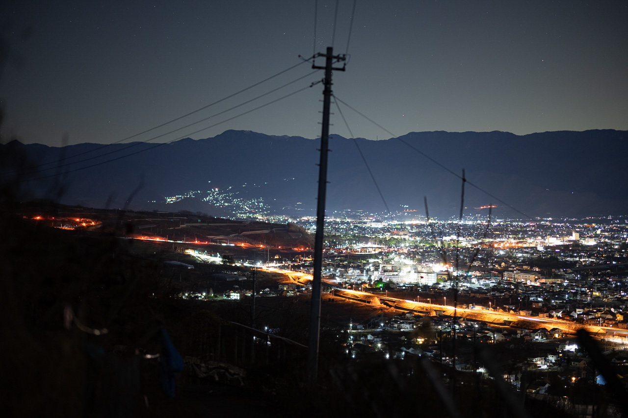 人の暮らしがある象徴としての夜景の上に、常に星空があるのだということを表現する作品。夜景を眺める場所が遠い場所にあるものではなく、生活圏の近くであることを意識させるものとして、電信柱を配している。また電信柱の先にはかに座を配し、星座の形にも視線が向くよう構成している。<br>Nikon Z 7 / NIKKOR Z 58mm f/0.95 S Noct / マニュアル露光（F0.95、0.8秒、-0.33EV） / ISO 1000