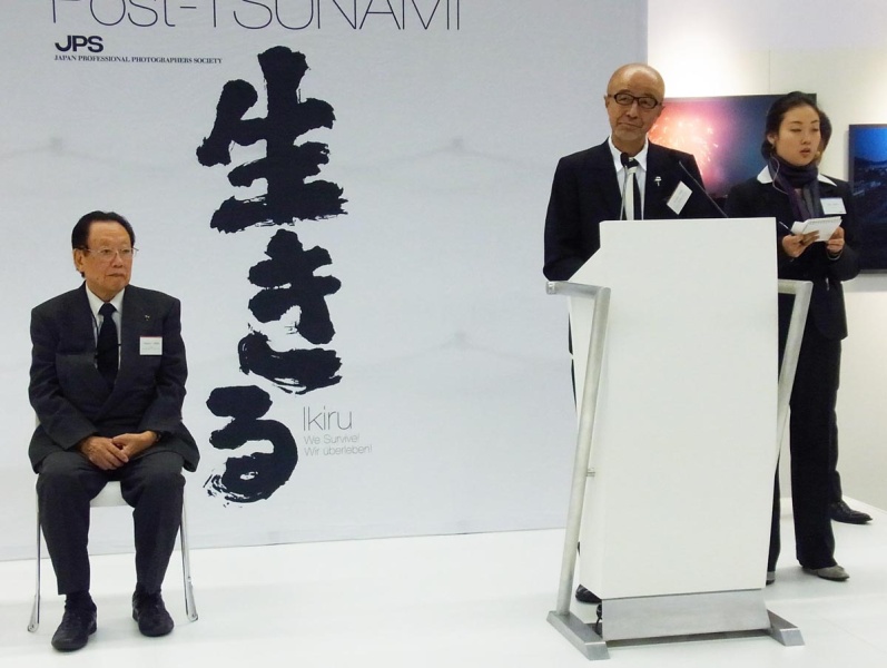 「生きる」写真展にて挨拶を述べる実行委員長の菅洋志氏と日本写真家協会会長の田沼武能氏（いずれも当時）、2012年フォトキナ会場にて。市川さん提供