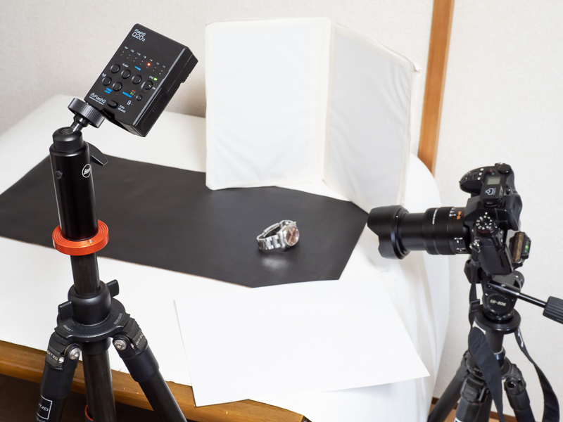 撮影の様子。Q20IIを三脚に装着し、時計の左斜め上から照射している。バウンスはさせず、直接光を当てることで精密機械が持つ金属的なイメージを強調した。右側と時計の左下には白レフを置いて明るさのバランスを取ると共に金属製のベルトに写り込ませてハイライトを作った。