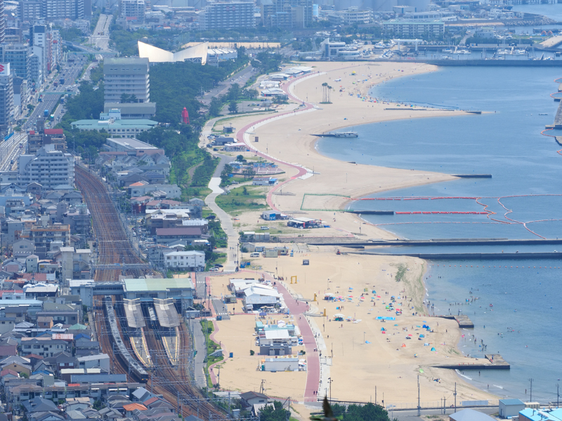 山陽本線の須磨駅を見下ろす。隣には駅直結型の須磨海水浴場が広がる。海風の影響を受ける中、手ぶれ補正の効果で構図を安定させ、列車の位置を見極めてシャッターを切った。<br><span class="fnt-85">OM-D E-M1X / M.ZUIKO DIGITAL ED 40-150mm F2.8 PRO / 106mm（212mm相当） / マニュアル露出（F10、1/500秒） / ISO 200 / WB:晴天</span>