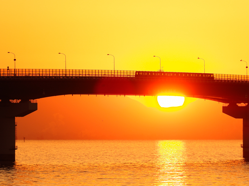 海上にできた神戸空港へアクセスするポートライナー。冬至前後の日の出を狙った。強烈な太陽光だが暗部が程よく保たれて列車も判別できる。背景のトーンも優秀だ。<br><span class="fnt-85">OM-D E-M1 Mark II /M.ZUIKO DIGITAL ED 40-150mm F2.8 PRO+Teleconverter MC-14 / 190mm（380mm相当） / マニュアル露出（F5.6、1/500秒） / ISO 200 / WB:日陰</span>