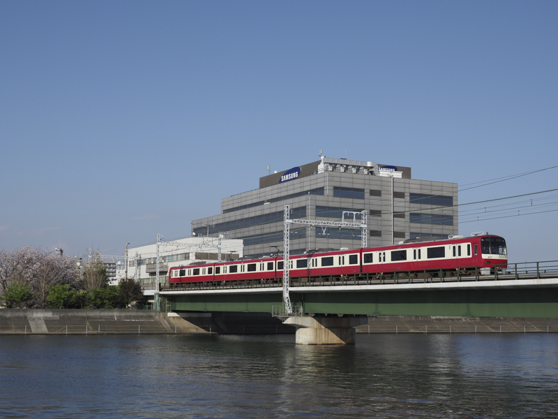 鶴見川を渡る京急線の1000形車両を順光で撮影した。「オリンパスブルー」の澄んだ青空と列車の赤色の組み合わせが心地良い<br><span class="fnt-85">OLYMPUS OM-D E-M1 Mark II / M.ZUIKO DIGITAL ED 12-100mm F4.0 IS PRO / 25mm（50mm相当） /絞り優先AE（F4、1/2,500秒、±0EV） / ISO 200 / WB:オート</span>