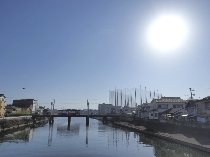 石津川の鉄橋を渡るかわいらしい車両。すでに中空高く昇った太陽と正対する画角。電車も背景に溶け込まずにきっちりと描写できた。小さなレンズがより愛おしくなる瞬間だ<br><span class="fnt-85">OLYMPUS OM-D E-M1 Mark III / M.ZUIKO DIGITAL ED 12mm F2.0 / 12mm（24mm相当） / 絞り優先AE（F7.1、1/1,000秒、-0.3EV） / ISO LOW（64相当） / WB:6,000K</span>