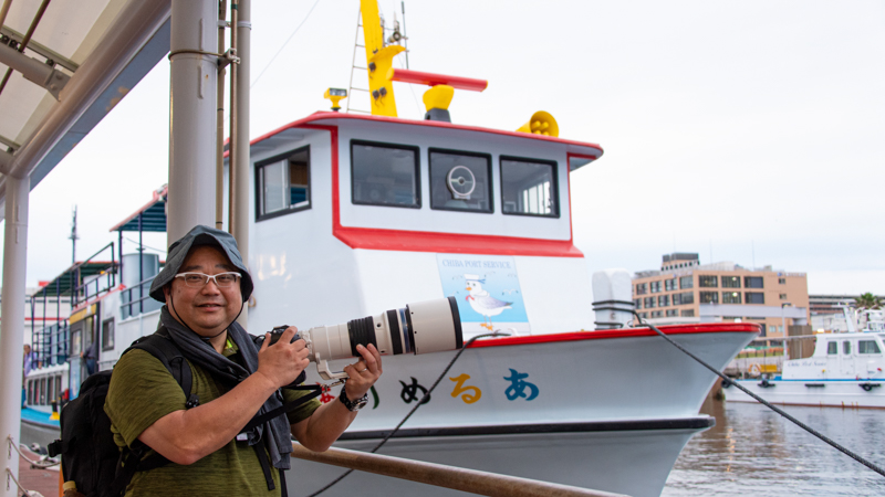出航直前の船の前にて。クルーズ船としては比較的小ぶりでカラフルな色使いが可愛らしい。この時点ではまだ、このあとの船の揺れの大きさも知らず、呑気な笑顔でいられたのだが……。（撮影：浅倉真一）