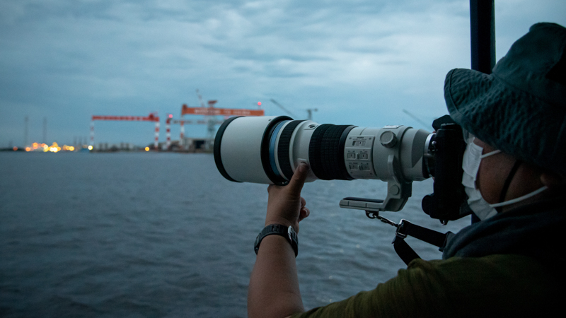 E-M1X + M.ZUIKO DIGITAL ED 150-400mm F4.5 TC1.25x IS PROで工場夜景を激写。カメラを構えた状態で全身をクッションのようにして揺れを吸収しつつ、ファインダーを覗きながら、像が安定した瞬間にシャッターを切る。船の揺れで想像以上にカメラが振られてしまうので、連写モードでとにかく数多く撮影しておくのがコツだった。なお低照度下のため、AF動作の逡巡はどうしても多くなる。あらかじめレンズのFnボタンにAF停止もしくはプリセットフォーカスを設定しておき、フォーカスが迷った際にはFnボタンでいったんフォーカス位置をリセットし、MFで微調整するのが効果的であった。（撮影：浅倉真一）