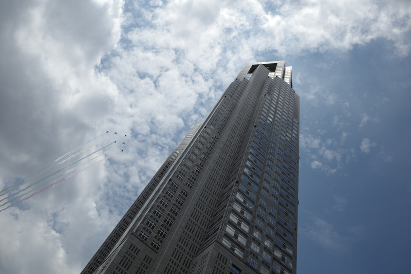 練馬方面から新宿上空を通過して都心に向かう、最初の飛来シーンです。都庁舎を射抜くイメージが狙いでしたが、想定よりもビルの頂上寄りを通過しそうだったので、とりあえず機体が都庁ビルの少し手前に来たところで1枚目を撮影。この直後に構図を少し右に振って修正し、フレーミングを固定した状態で機体が都庁の右側に抜けるのを待ちました。<br>RICOH GR III（28mm相当）シャッター速度優先AE・1/1,000秒・+1/3EV・ISO 100・AWB