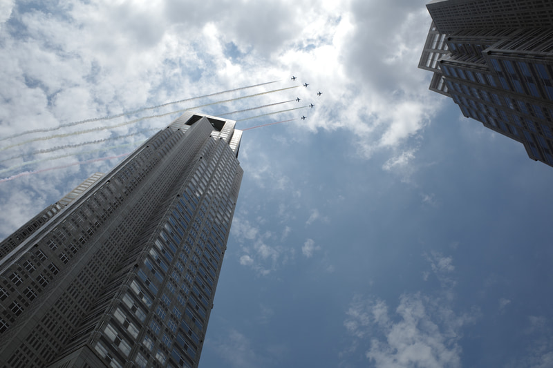 待ち伏せて連写したこの1枚目が、最初の飛来シーンでは結果的にベストのタイミングとなりました。というのも、この直後にスモークがオフになったからです。太陽光を受けた明るい雲が画面内にあったので、+1/3の露出補正で画面全体が暗くならないよう設定しています。過度のプラス補正はカラースモークの濃度も薄くなるので注意が必要です。ビルの間を抜けていくイメージに構図変更したつもりでしたが、少々中途半端になってしまいました。前のカットも同様ですが、真四角（1：1）にトリミングするのもありかと思います。そのようなトリミングの自由度という点でも、望遠より広角のレンズが少し有利と言えます。<br>RICOH GR III（28mm相当）シャッター速度優先AE・1/1,000秒・+1/3EV・ISO 100・AWB