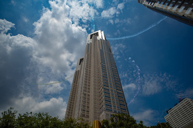 さらに約4分後、最後は五輪マークを描く飛行です。展示エリアは新宿エリアの真上だったため、スモークが都庁舎に遮られる部分もあり、予想した通りマークの全体像を見渡すことはできませんでした。それでも円を描いて飛ぶ機体とスモークが複数写るように、数枚撮影してみました。スモークを引く機影が3つと、スモークを出さないリーダー機がかろうじて写っています。<br>RICOH GR III（28mm相当）シャッター速度優先AE・1/1,000秒・+1/3EV・ISO 100・AWB（PhotoshopでRAW現像）