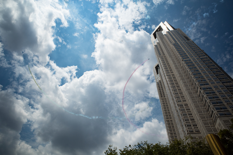 撮影対象を緑と赤のスモークを引く2機に絞り、カメラを左に振って4枚連写。その3枚目の写真です。スモークがなるべく長く写ることを意識しましたが、この次の4枚目では赤いスモークを引く機体が都庁ビルに近づきすぎてしまったため、このカットを選択しました<br>RICOH GR III（28mm相当）シャッター速度優先AE・1/1,000秒・+1/3EV・ISO 100・AWB（PhotoshopでRAW現像）