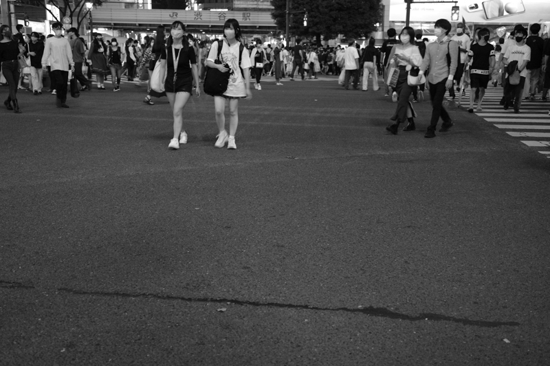 渋谷のスクランブル交差点で信号待ちをする女性。マスクとスマホとワイヤレスイヤフォン、それが今という時代の定番<br><span class="fnt-85">RICOH GR IIIx／40mm／プログラムAE（F2.8、1/40秒、－0.7EV）／ISO 1000／オートWB／モノトーン</span>