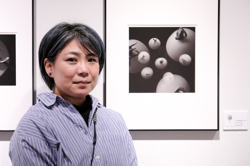 こばやしかをるさん（「curiosity —好奇心—」会場にて）。向かって右奥の展示作品は国際写真賞IPA 2021のプロフェッショナル ファインアート・静物部門で佳作に入選した作品。当誌で2020年5月にお伝えしたRICOH GR IIIを用いた多重露光機能による室内撮影に着想を得て取り組んだ結果生まれた作品なのだという。
