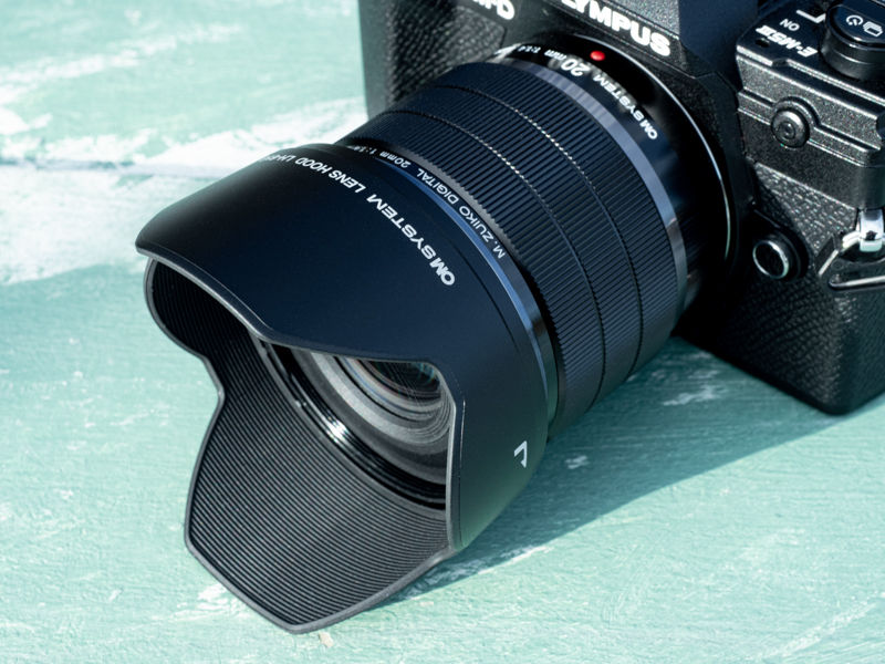 同梱のレンズフード「LH-61G」を装着。鏡筒のサイズから考えると比較的大きめなイメージとなる。取り付け部にロック機構はない。本製品からブランド名がOLYMPUSからOM SYSTEMに変更されており、レンズ鏡筒だけでなく、フードやキャップのロゴも刷新されている。