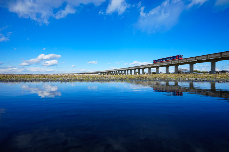 鹿島臨海鉄道のベストショットはこちら！ やはりこの路線の特徴である「単線・非電化・高架」区間が主役ですが、雨の翌日に道路にできた水たまりを使って撮影することで、まるでウユニ塩湖で撮影したかのような、幻想的な風景になりました。行ったことないけど（笑）。<br>ソニーα1　FE 16-35mm F2.8 GM（16mm）　絞り優先オート　（F16、1/500秒）　ISO 800　WB:太陽光　<a href="https://goo.gl/maps/uXcNAHFFx1pTkBZW6" class="n" target="_blank">[Google Maps]</a>