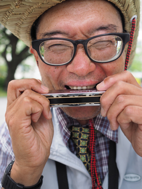 街のハーモニカ吹きさん。若い時に高齢者問題のルポの仕事をしたことがあります。この時は「老人写真家」と呼ばれたこともありましたが、自分が年寄りになりますと仲間意識が芽生えて撮影したくなります。絞りを開いてカメラ任せの顔認識AFで撮影しました。ファインダーはろくに見てませんが、きちんと目に合焦します。<br>PEN-F　M.ZUIKO DIGITAL 17mm F1.8（F2.2・1/800秒）ISO 400