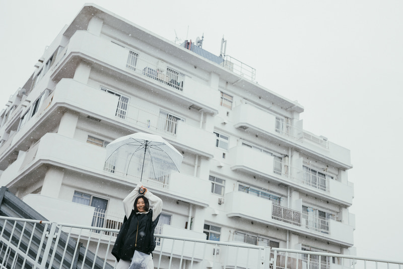 ライカSL2-S／アポ・ズミクロンSL f2/35mm ASPH.／35mm／マニュアル露出（F2、1/800秒）／ISO 100／WB：5,300K<br>少し坂をのぼった先にあったマンション。パッと見た瞬間、ローアングルで撮りたいと思ったが、彼女の存在感は薄れてしまう。「傘を両手で高く持ち上げてほしい！」 こんな傘の持ち方なんて日常ではしないけど、だからこそ人は笑顔になる