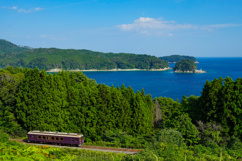もとJR山田線の区間である岩手船越駅と浪板海岸駅の間のポイントです。震災後に三陸沿岸道路が建設され、その工事用の道路から何箇所か海と列車が撮影できるようになりました。ここではカットしていますが、写真のすぐ下は高速道路という状況になっています。ただ三陸鉄道リアス線全体を見ても、海と列車を防風壁などの邪魔なものなしで撮影できる貴重なポイントと言えるでしょう。ただし年々木々が伸び、いつ撮れなくなってもおかしくない状況ですので、撮影するならお早めにどうぞ。<br>ソニーα1　FE 24-70mm F2.8 GM（37mm）マニュアル露出（F8、1/800秒）ISO 800 WB:太陽光　<a href="https://goo.gl/maps/9GDdRZtmaboQjzeUA" class="n" target="_blank">[Googleマップ]</a>