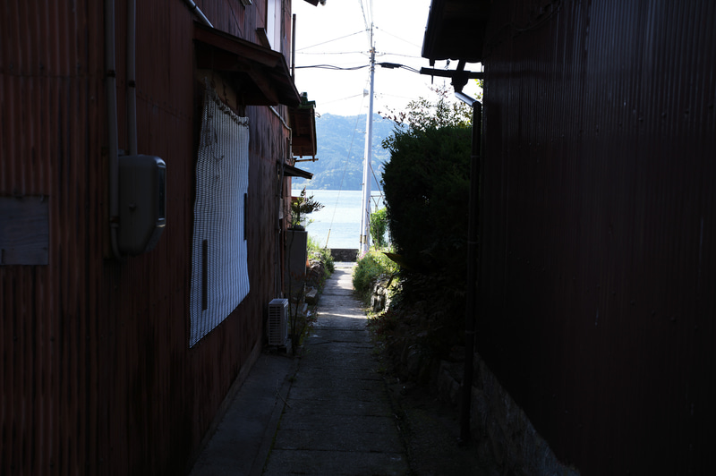 沖島は琵琶湖に面するメインストリート以外、細い路地が家と家の間を通っている。そこから見る琵琶湖は、いつもの雄大さが影を潜め、まるで遠い世界をのぞき見ているような感覚に陥ってしまう。<br>ライカM11　アポ・ズミクロンM f2.0/35mm ASPH.（F5.6、 1/160秒）ISO 125　WB:オート