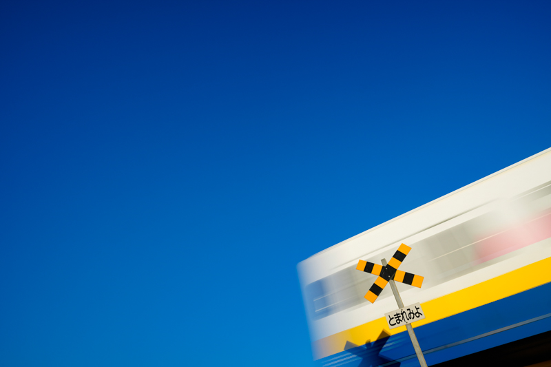 田んぼの中にある踏切は、警報機も遮断器もない「第4種踏切」です。周囲に高い建物などもないので、ローアングルで狙うと、青空バックで踏切を撮影できます。列車をそのまま写すと、列車と踏切が重なって目立たなくなってしまうので、1/15秒で列車をブラしてみました。列車の晴れた空の青と、列車の黄色いラインが、どことなくウクライナカラーを彷彿させ、お気に入りの作品になっています。<br>ソニーα1　FE 16-35mm F2.8 GM（23mm） 絞り優先オート （F5.6、1/15秒）ISO 100　WB：日陰　<a href="https://goo.gl/maps/pQDqfLS1fhG2RJFq6" class="n" target="_blank">[Google Maps]</a>