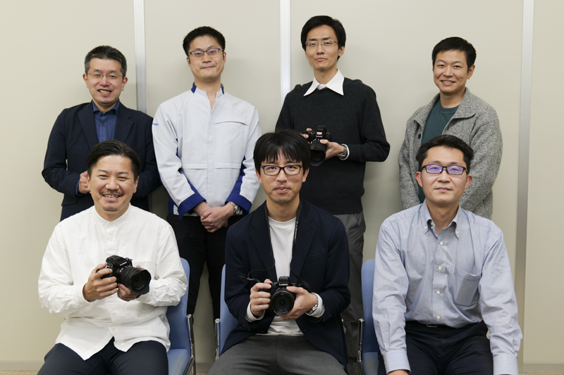 上段左から、櫻井幹夫氏（手ブレ補正）、金田憲和氏（外装設計）、栃尾貴之氏（画質）、塩見記章氏（マーケティング）、渡邊慎治氏（商品企画）、中村光崇氏（開発リーダー）、大神智洋氏（AF）<span class="fnt-85">※かっこ内は主な担当分野</span>