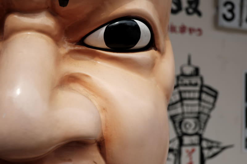 串揚げ屋さんの看板人形ですけど、ただ撮影しただけではつまらない写真になってしまったので目玉だけをトリミングしてみましたが、ビクともしません。高画素機のフレキシビリティを見た思いです。<br>X-T5／XF18mmF2 R／絞り優先オート（F5.6・1/1,250秒）／ISO400