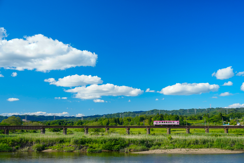 山形鉄道フラワー長井線で最近発見した、梨郷駅と西大塚駅の間にあるこの鉄橋。歴史ある切石積みの橋桁が素朴な雰囲気で最高です。青空にぽっかりと浮かぶ白い雲を見ながら走る列車は、まるでイラストのよう。山形鉄道の車両には沿線の花にちなんだラッピングが施されており、このピンク色の車両は南陽市のさくらがモチーフです。<br>ソニーα1／FE 24-70mm F2.8 GM II／絞り優先オート（38mm、F5.6、1/400秒）／ISO400／WB:太陽光　<a href="https://goo.gl/maps/uB76AkUS2vgxcM4G7" class="n" target="_blank">[Google Maps]</a>