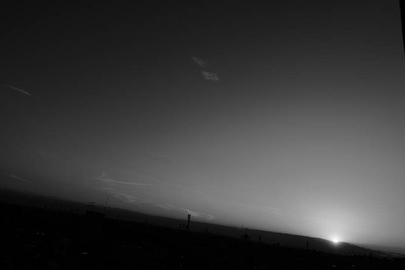 街の向こうに沈んでいく夕日。28mmで切り取る自然な画角が好きだ。雲の中に入っていく太陽の輪郭が浮かび上がるまで露出を抑えている。<br>ライカSL2-S／アポ・ズミクロンSL f2/28mm ASPH.／プログラムAE（F5、1/1,000秒、-1.7EV）／ISO 100／WB：オート／モノクロHC