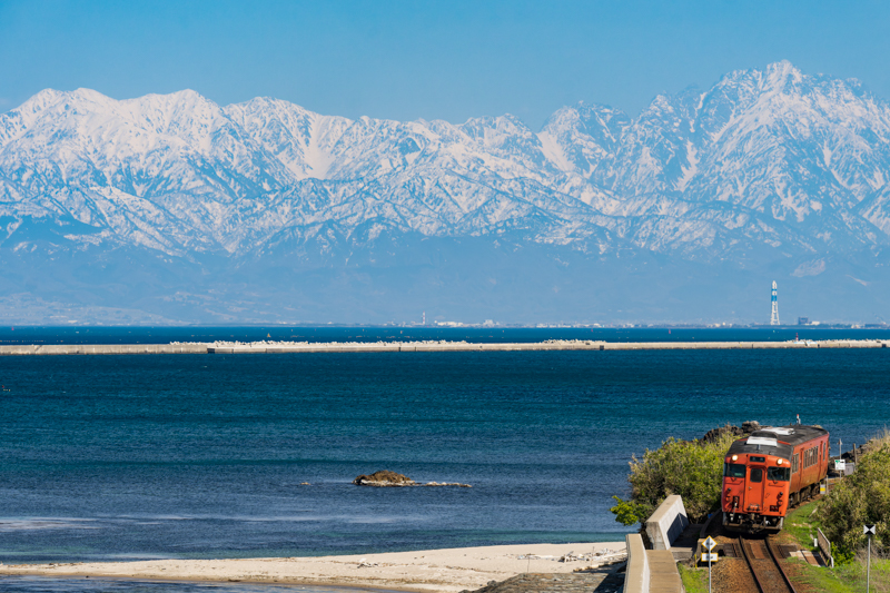 富山湾の奥に広がるのは勇壮な立山連峰。その横を氷見線が走る風景は、まさに富山を代表する絶景でしょう。こんなにも素晴らしい風景ですが、撮影ポイントは道の駅「雨晴」の展望台。美味しい食事やスイーツも楽しめる、天国のような撮影地なのです。ただし作品ではギリギリカットしていますが、画面右側すぐのところに国道が並走しているので、構図は限られてしまいます。展望スペースは三脚禁止ではありませんが、観光客もたくさん来る場所なので、できれば手持ちで、長時間の場所確保は避けたいところです。<br>ソニーα1／FE 70-200mm F2.8 GM OSS II／絞り優先AE（191mm・F8・1/500秒）／ISO 400／WB:太陽光　<a href="https://goo.gl/maps/NffQV4i9wi8qAEpK9" class="n" target="_blank">[Google Maps]</a>