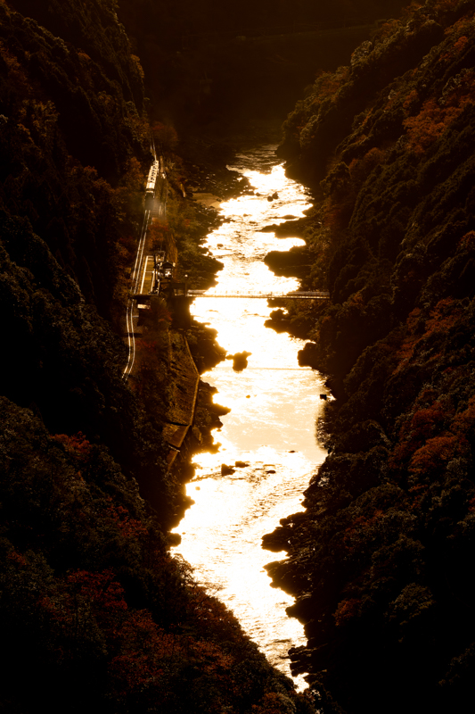 「トロッコ保津峡駅」と保津峡を大俯瞰できる、とっておきのポイントです。この作品は12月の15時ごろに撮影したのですが、ちょうど川の真上に来る太陽を受けて保津峡がギラギラと輝き、息を飲むほど幻想的な光景になりました。トロッコ列車の屋根が、まるで僕の青春時代に乗っていた山陰本線の旧型客車みたいに輝き、ちょっと感動してしまいました。<br>実はこの写真、有料道路「嵐山-高雄パークウェイ」にあるビュースポット「保津峡展望台」からの撮影で、徒歩やバスなどの公共交通では行くことができません。有料道路のため通行料金（軽・普通自動車）が1,200円必要となりますので、ここぞというタイミングで訪ねるといいでしょう。<br>ソニーα1／FE 70-200mm F2.8 GM OSS ／絞り優先オート（176mm・F11・1/800秒）／ISO 200／WB:日陰 <a href="https://goo.gl/maps/C4EStt37tDKpRBgr5" class="n" target="_blank">[Google Maps]</a>