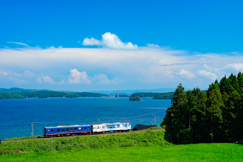 のと鉄道は能登半島の海沿いを走りますが、蛸島までの路線が健在だったころから、海と列車をからめて撮影するのが難しい路線でした。そんななか、海と列車を撮影するオススメのポイントがこちら。段々畑の上にある山の斜面から見下ろすと、海に浮かぶ能登島と、「ツインブリッジのと」を入れた絶景を撮影することができます。順光になる午後がオススメです<br>ソニーα1／FE 24-70mm F2.8 GM／絞り優先オート（47mm・F8・1/800秒）／ISO 1000／WB:太陽光／<a href="https://goo.gl/maps/nT31WrRXwyZokgFp9" class="n" target="_blank">Google Maps</a>