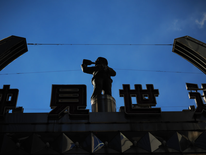 商店街のアーチの上に酔っ払いのおっさんが逆光で照らされているのを発見したので何も考えずに撮影と。よく写りますねー<br>OM-1／M.ZUIKO DIGITAL ED 30mm F3.5 Macro／絞り優先AE（F4・1/5,000秒・-1.3EV）／ISO 200