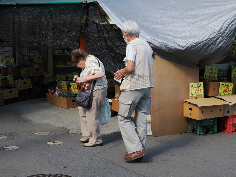 八百屋さんで買い物中の老夫婦です。すでに筆者も仲間意識があるのか、つい撮影をしてしまいまして。背景の日除が、舞台装置の幕みたいでいいなと<br>OM-1／M.ZUIKO DIGITAL ED 30mm F3.5 Macro／プログラムAE（F8・1/320秒・-0.7EV）／ISO 400