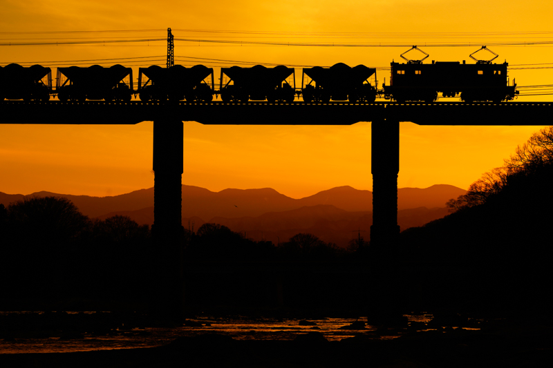 上長瀞駅〜親鼻駅間に位置する荒川橋りょうは、秩父鉄道を代表する撮影地。私鉄では今や貴重な存在となった貨物列車をシルエットにして撮影しました。こういう撮影では、列車の足回りまでしっかりと影絵になっていることが大切。鉄橋の撮影では、どうしても鉄橋下から見上げたくなりますが、それだと列車の車輪が鉄橋と同化してしまい美しいシルエットになりません。ここではあえて鉄橋から離れて望遠レンズで狙うことで、車両の隅々まで精緻に影絵にすることができました。シルエット撮影は列車の色や質感を表現できませんが、影絵にすることで、かえってフォルムの美しさが強調されるから不思議ですね<br>ソニーα7R III／FE 100-400mm F4.5-5.6 GM OSS／マニュアル露出（146mm相当・F9・1/1,000秒）／ISO 200／WB:日陰　<a href="https://maps.app.goo.gl/NdKn2fzsydTj8QQm8" class="n" target="_blank">[Google Maps]</a>