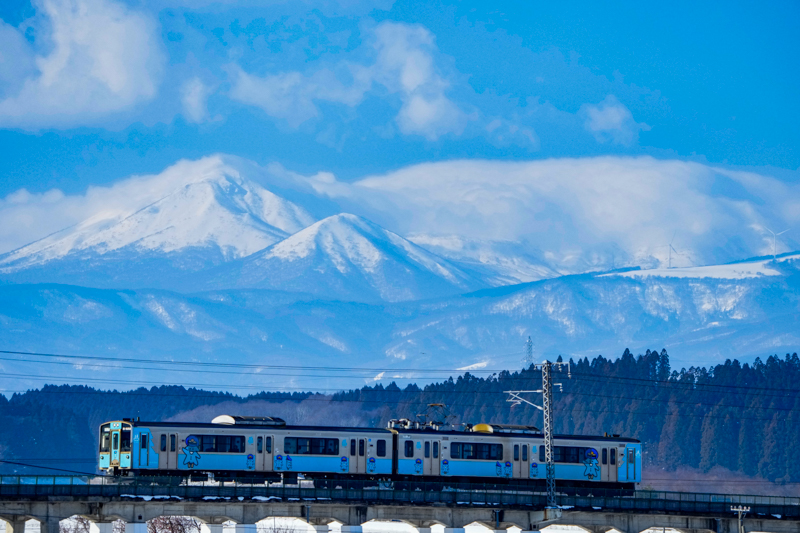 こちらは青い森鉄道と八甲田の山々をからめて撮影できるポイントです。八甲田連峰は主峰である大岳を中心とした山々の総称ですが、かなり奥まった位置にあり横に長く広がっているので、列車が走る位置からだと、岩木山のように明快に撮ることが難しい山であるように感じます。ただしこのポイントでは、望遠レンズで雪をたたえた山々を圧縮できるため、迫力ある八甲田連峰の山容と列車をからめて撮れる貴重なポイントといえるでしょう。終日逆光気味なので、日が当たる午前中早い時間帯が狙い目です（青い森鉄道 上北町〜小川原）<br>ソニーα1／FE 200-600mm F5.6-6.3 G OSS／絞り優先オート（321mm・F6.3・1/2,500秒）／ISO 1600／WB:太陽光　<a href="https://maps.app.goo.gl/vwK2fH4ZSzpRm3MW8" class="n" target="_blank">[Google Maps]</a>