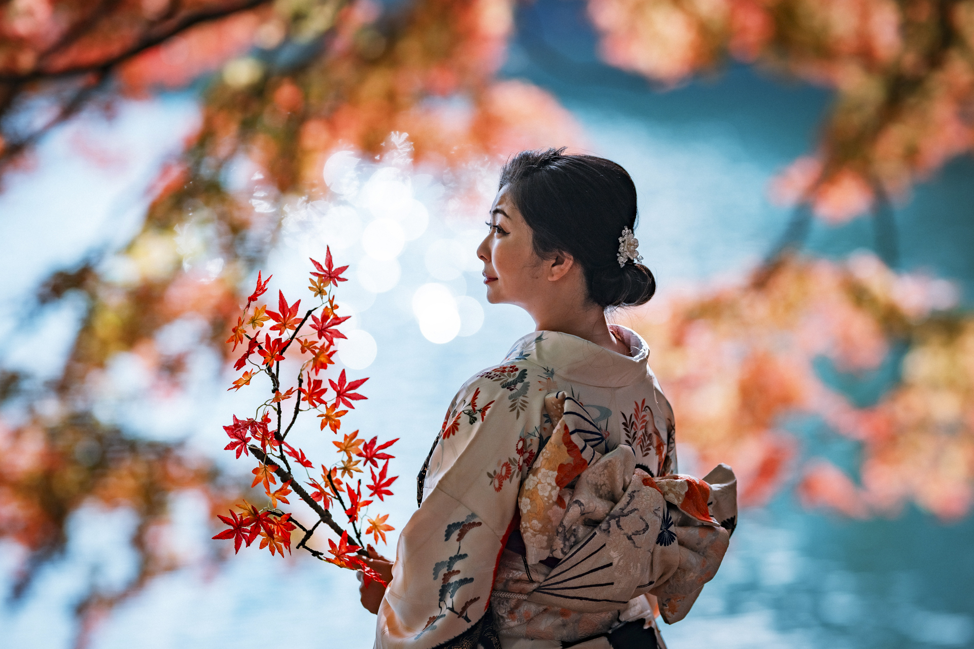 秋の湖。水面に強い太陽の光が反射してキラキラと光る逆光のシーン。水面のキラキラがきれいな丸ボケの背景となって人物が立体的に浮かび上がった。モデルが手にしているのはリアルな造花<br><span class="fnt-85">Z 9／NIKKOR Z 135mm f/1.8 S Plena／マニュアル露出（1/4,000秒、F1.8）／ISO 160</span>