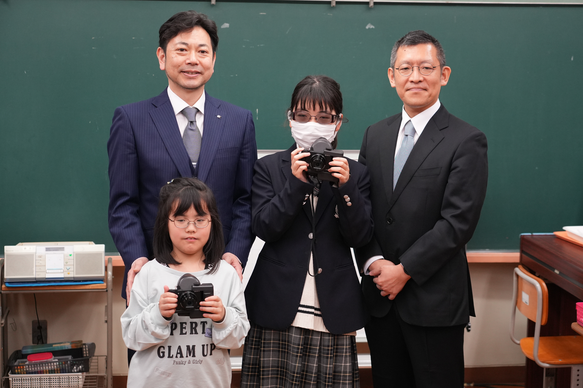 東京都立八王子盲学校で行われたカメラの寄贈式にて。後列左がソニー株式会社イメージングエンタテインメント事業部・大島正昭事業部長、後列右が株式会社QDレーザ・宮内洋宜事業推進室室長、前列は八王子盲学校の生徒2名