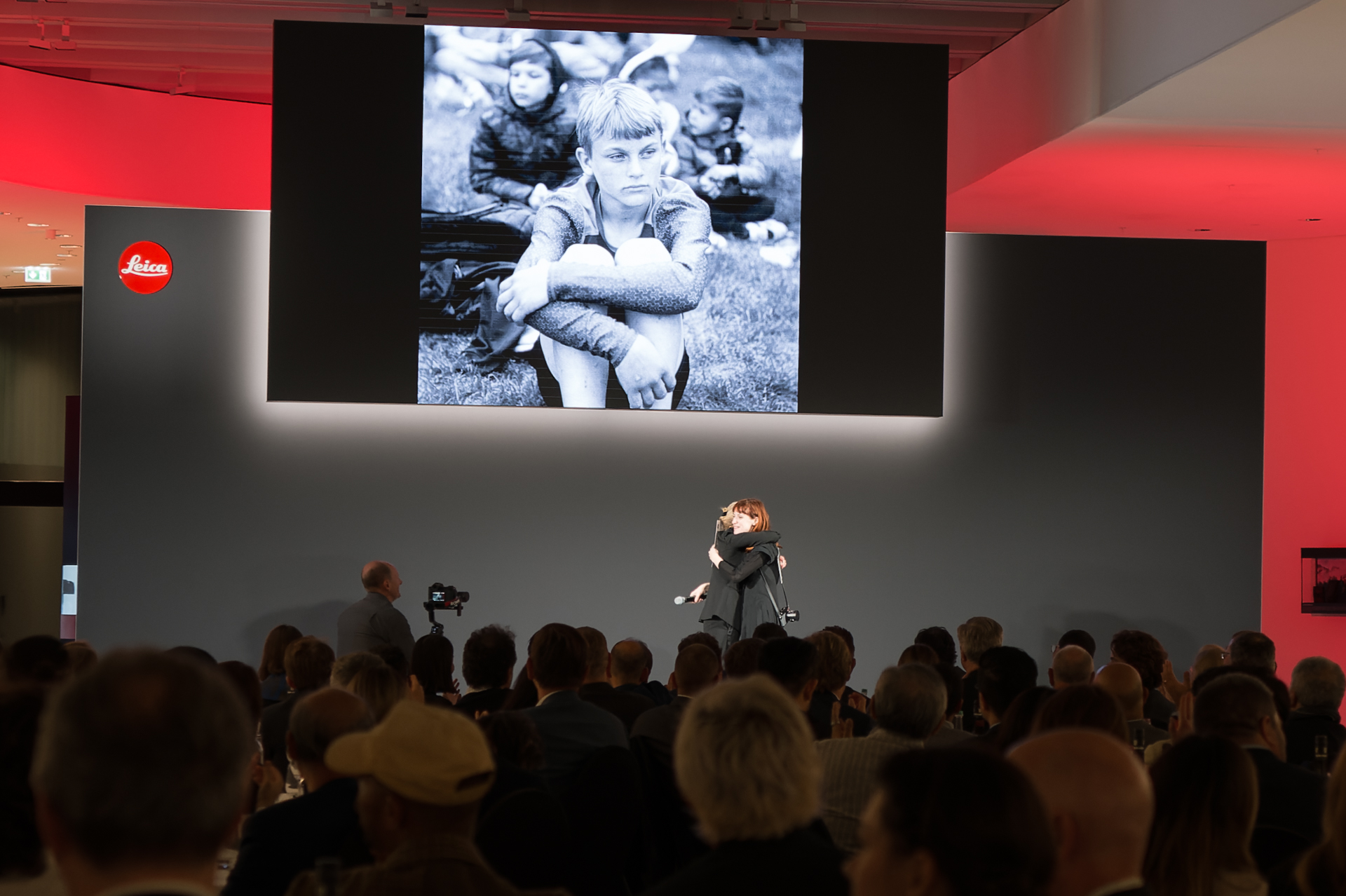 贈られたライカQ3（張り革の色から「ライカQ3 43」と見られる）を受け取り、カリン・レーン=カウフマン氏とハグ