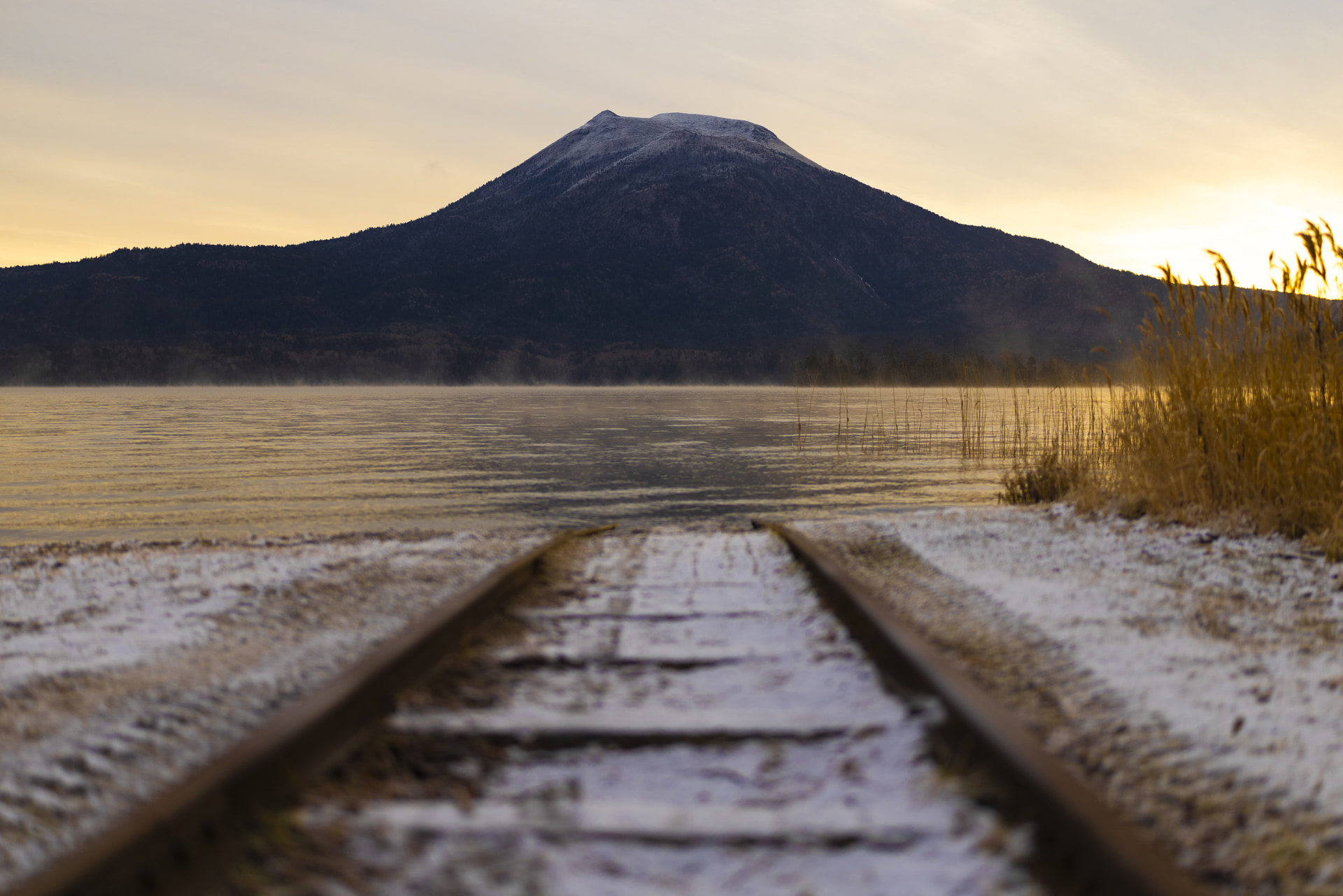 気嵐（けあらし）が立つ阿寒湖の朝。目の前にそびえる雄阿寒岳にフォーカスした。開放絞りのF1.4でローポジションから撮影することで、湖に向かって伸びるレールを前ボケとして表現し、写真に奥行きを生み出した<br><span class="fnt-85">キヤノン EOS R5 Mark II／RF50mm F1.4 L VCM／50mm／マニュアル露出（F1.4、1/1,250秒）／ISO 50／WB：オート</span>