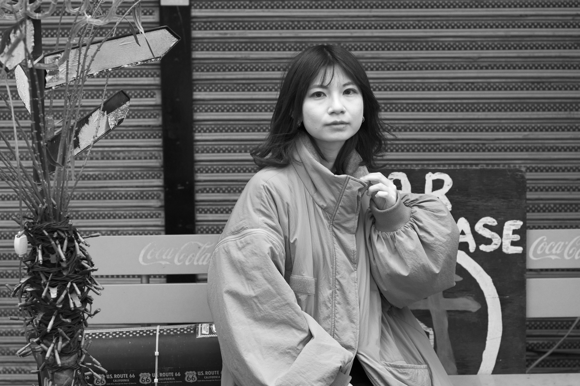 背景のシャッターは青色、ベンチは赤色というサイケ感が個人的に気に食わずピクチャーコントロールでディープトーンモノクローム設定。この条件ではフツーのモノクロの調子ですね。モデル：<a href="https://x.com/okw_hi_">ひぃな</a><br><span class="fnt-85">ニコン Z50II／NIKKOR Z 50mm f/1.8 S／50mm（75mm相当）／絞り優先AE（1/1,250秒、F2.5、−0.7EV）／ISO 400</span>